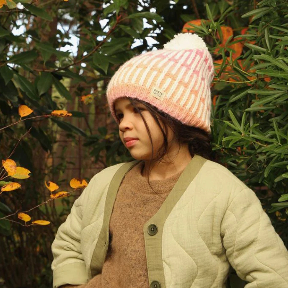 A child wearing a pink striped beanie hat with a pom-pom, standing in front of foliage.
