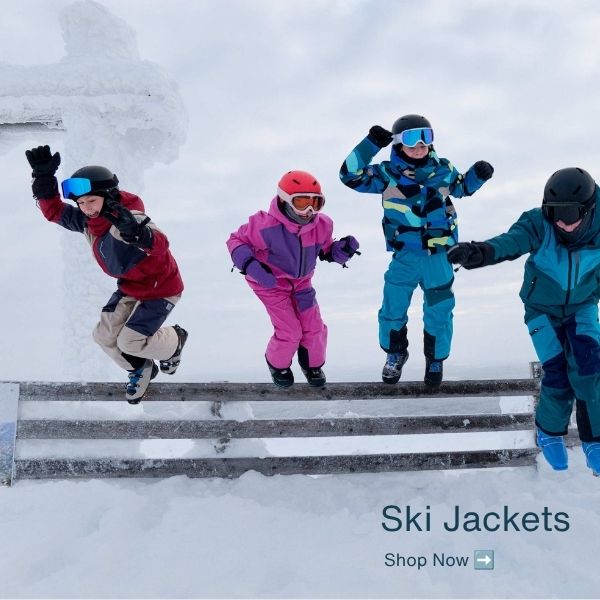 Four children in colourful ski jackets and snow gear jump over a snowy barrier at a ski resort, with the text 'Ski Jackets' and 'Shop Now' in the lower right corner.
