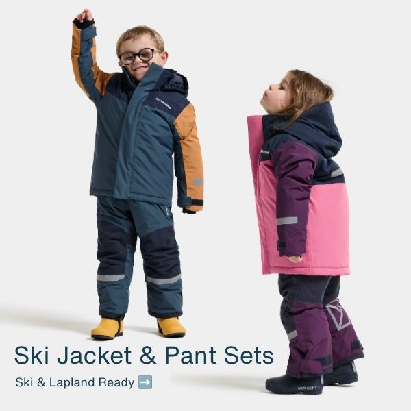 Two children modeling winter ski jackets and pant sets against a light gray studio background. The child on the left wears a navy and tan jacket with navy pants and yellow boots, while the child on the right wears a pink and navy jacket with dark purple pants.